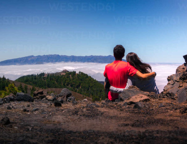 Back view of tender traveling couple sitting on hill and hugging while admiring spectacular scenery on clouds over mountainous terrain